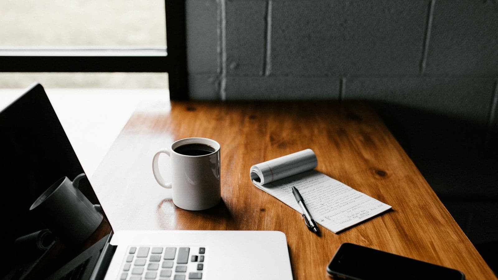 A desk with a laptop, a coffee cup and a notepad of handwritten notes.